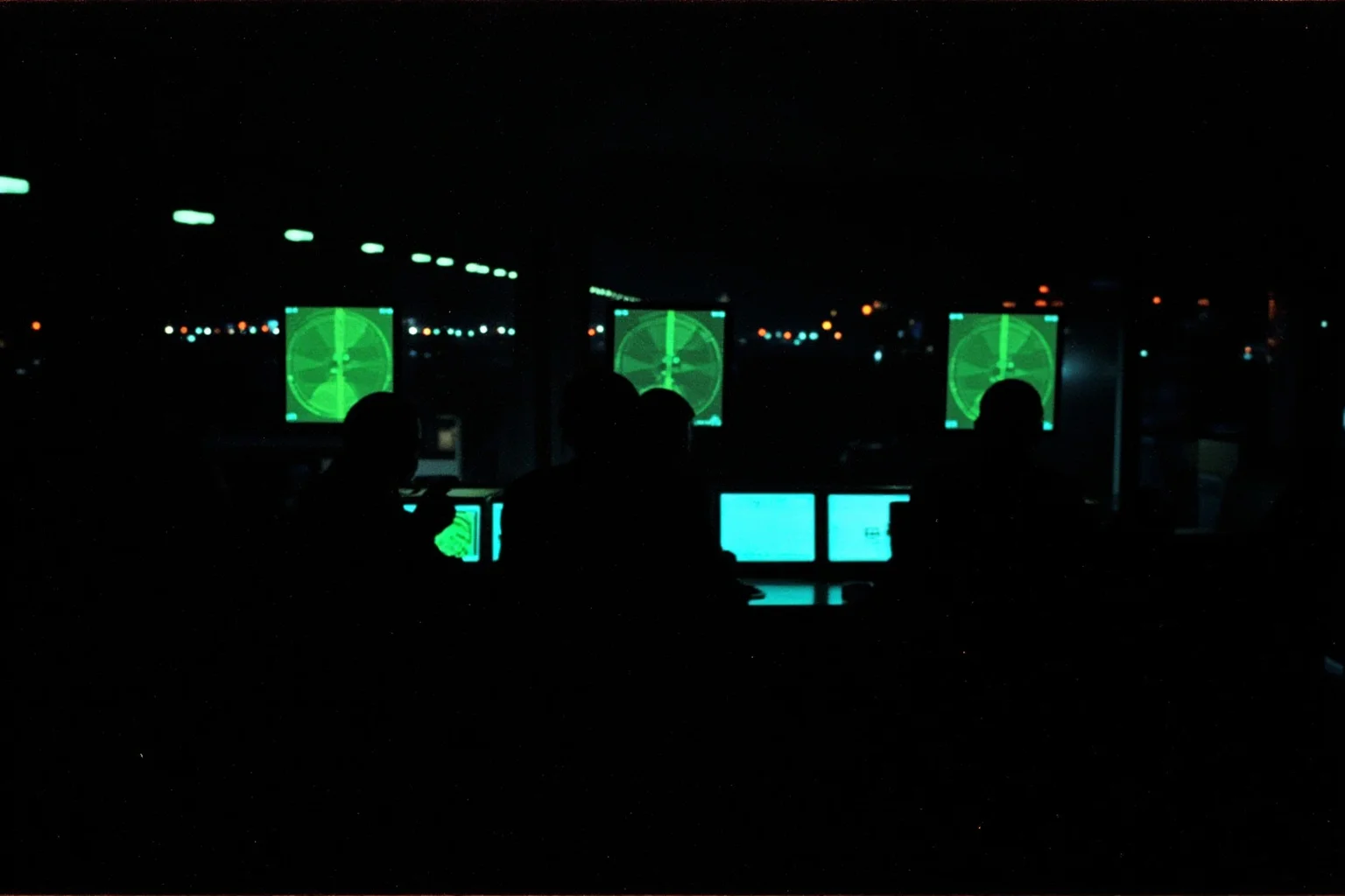 Air traffic control tower interior at night in the late 1990s with green radar screens, silhouetted controllers, and airport lights in the background