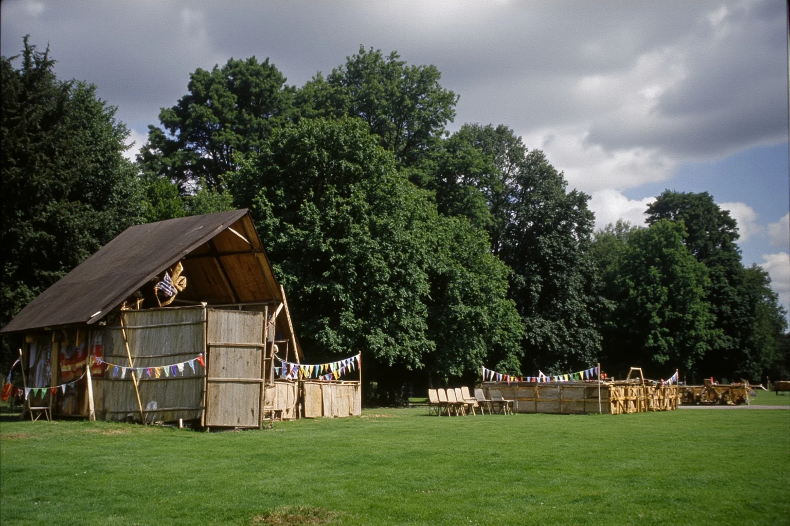 An English village green with a makeshift outdoor stage, folding chairs, bunting and darkening clouds on a summer afternoon, 1930s setting.