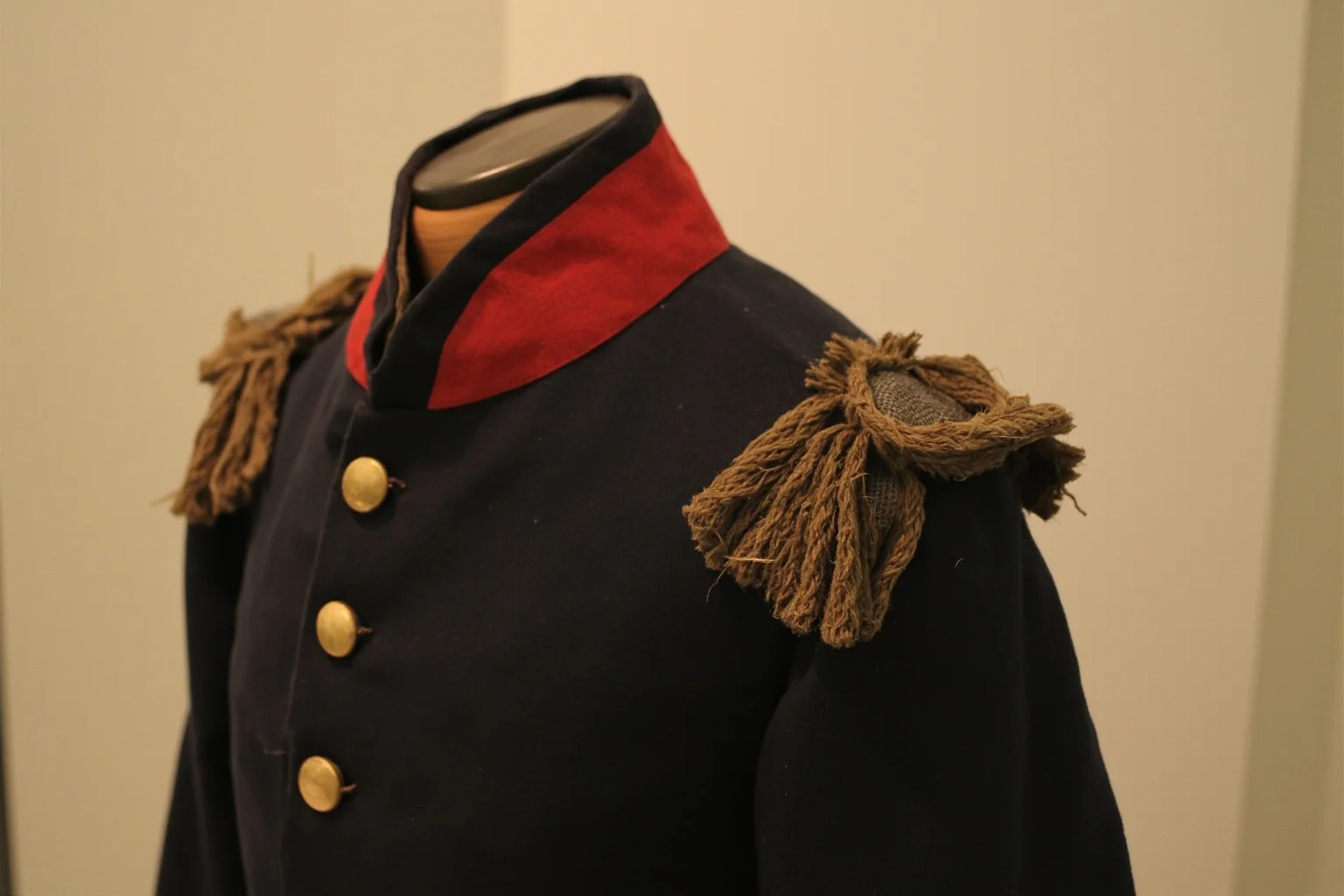 An old colonial military coat with a visible patched tear near the shoulder, displayed on a mannequin in a museum, no people.