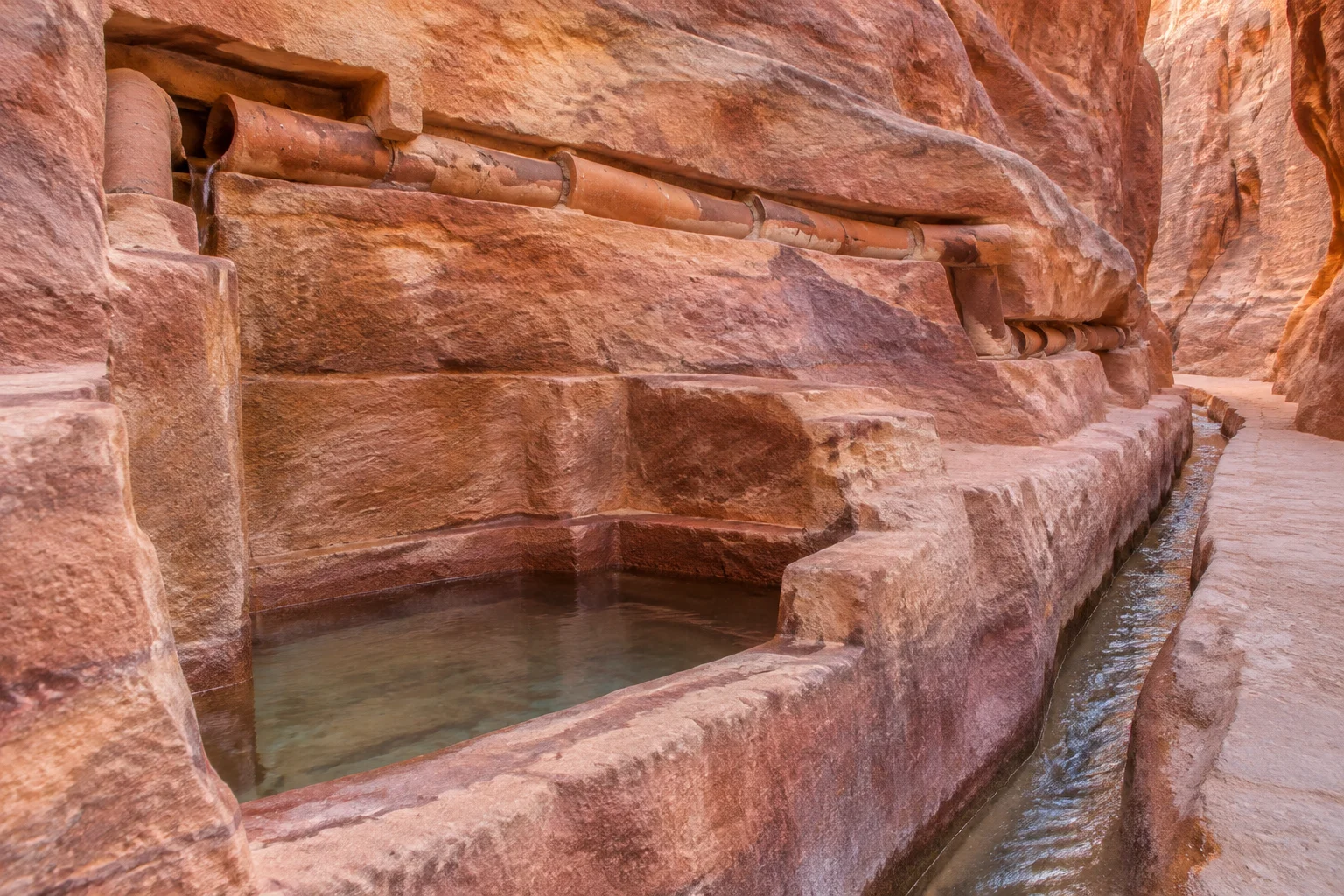 23 Historical Finds That Reveal Advanced Ancient Civilizations 7 Nabataean cistern and ceramic pipeline carved into the sandstone walls of the Siq at Petra in Jordan, water channel running along the rock face, warm rose-coloured stone, no people.