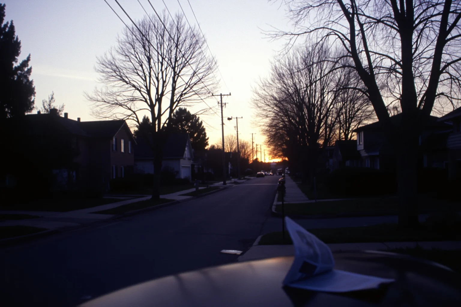 Sunrise over a quiet suburban street on the morning of January 1, 2000, porch lights still on, newspaper on the driveway, calm pale light