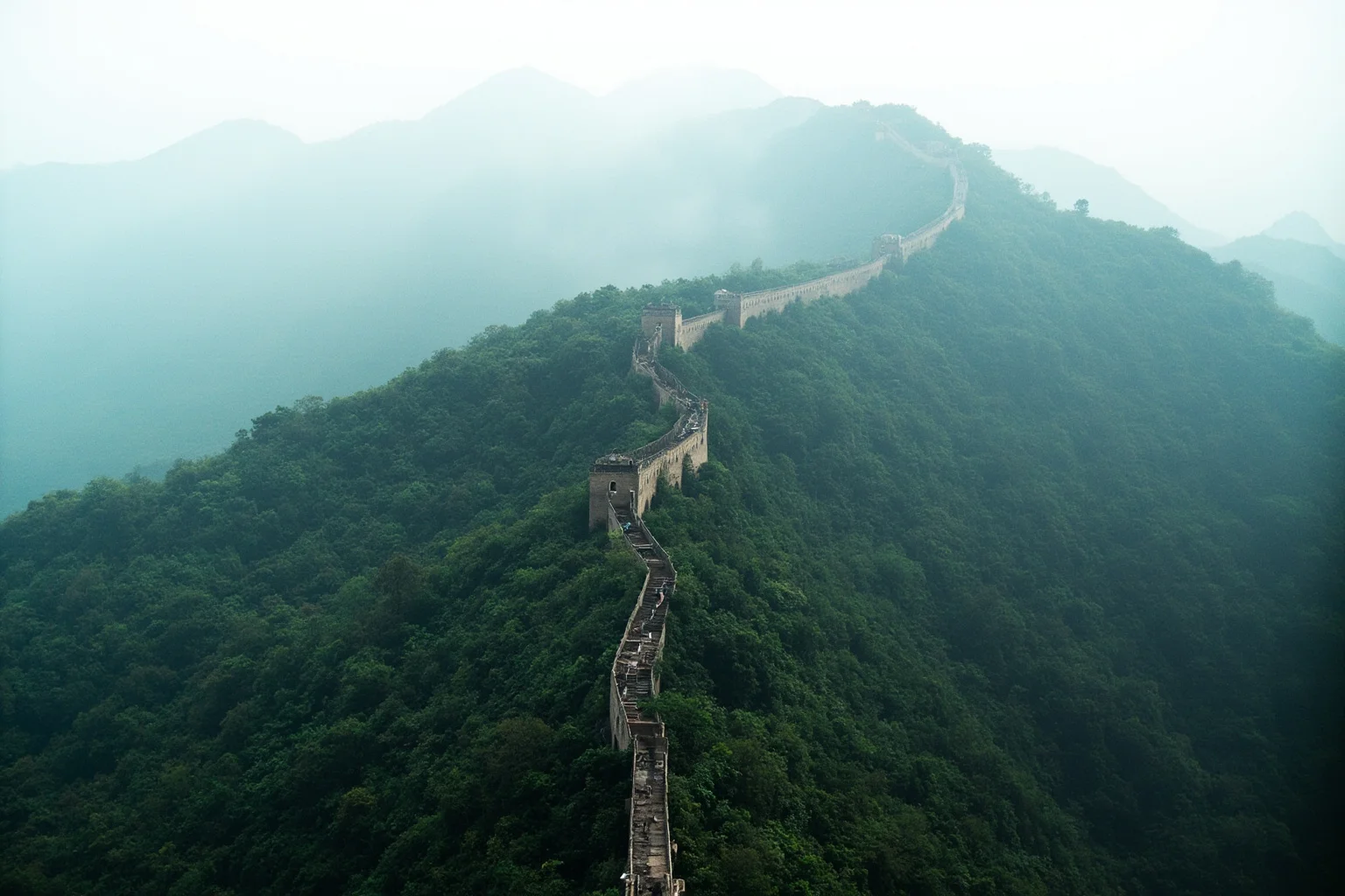 The Great Wall of China snaking over forested ridgelines in drifting mist, restored Ming-era brickwork