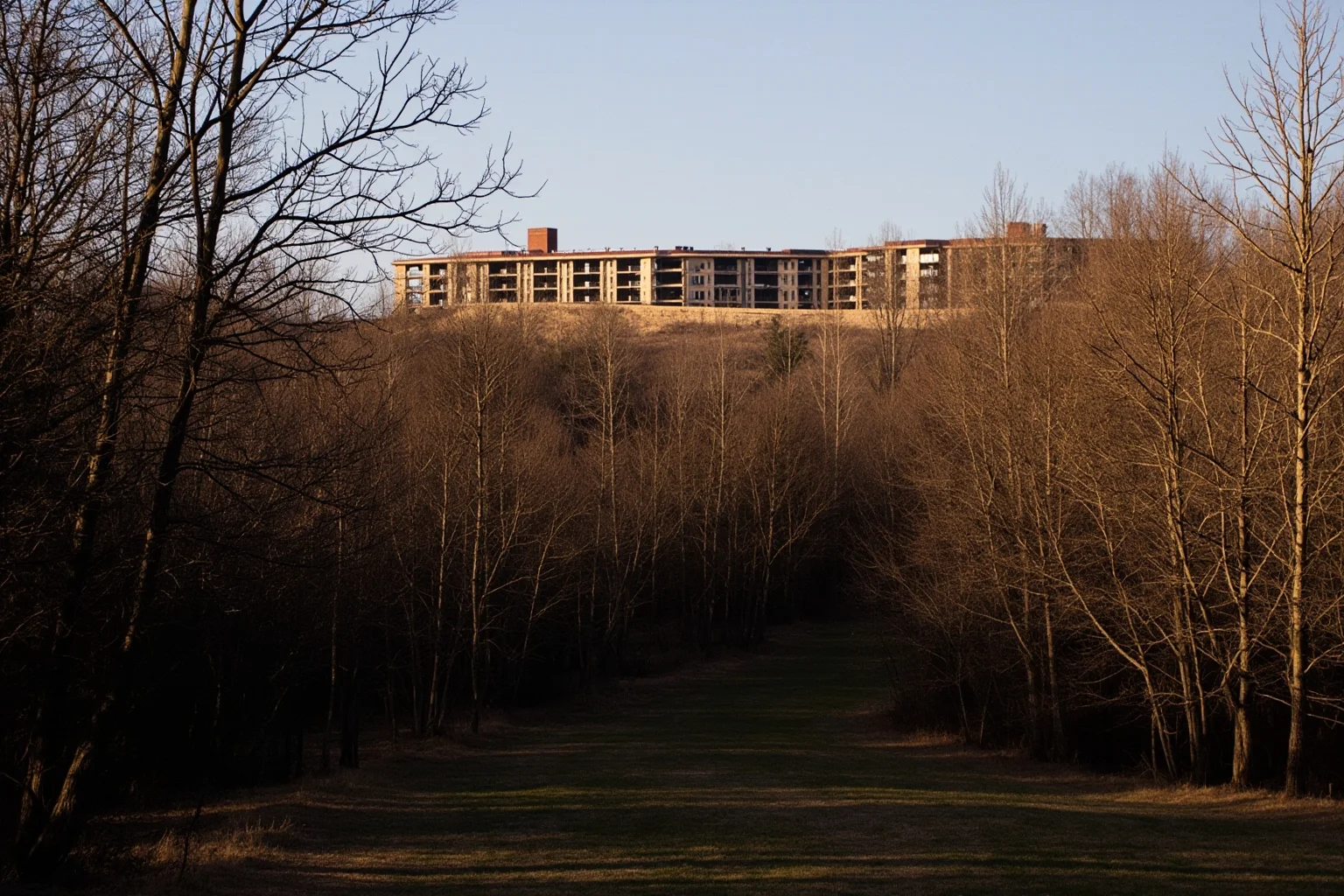 Wooded ridge with a large abandoned hospital complex behind trees, walking trail in foreground, golden hour, no people, photorealistic.